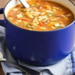 A steaming bowl of vegetable soup with colorful vegetables and herbs, served with a slice of crusty bread.